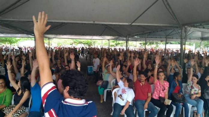 fim da greve em florianopolis centenas de pessoas sentadas em assembleia com o braço estendido para o alto; em primeiro plano, um homem de costas segurando um microfone com a mão para o alto