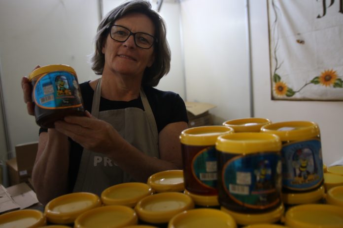florianopolis_-_feira_reune_produtores_catarinenses_de_mel_20180613_1928614048 em frente a uma bancada com vários potes de mel, mulher segura um pote e posa para câmera