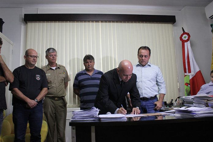 Homem careca se abaixa para assinar um papel sobre uma mesa, circundado por outros homens de pé com uma cortina ao fundo, em uma sala de gabinete