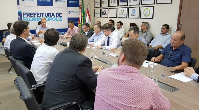 Vários homens reunidos em uma grande mesa com o prefeito Gean Loureuiro na ponta gesticulando enquanto fala. Ao fundo, um painel com logotipos da prefeitura e uma parede com quadros