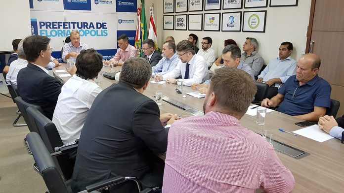 Vários homens reunidos em uma grande mesa com o prefeito Gean Loureuiro na ponta gesticulando enquanto fala. Ao fundo, um painel com logotipos da prefeitura e uma parede com quadros