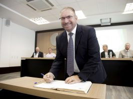 Prefeito Morastoni é eleito por aclamação na Fecam Morastoni, com uma caneta na mão e atrás de uma mesa com um papel no meio, sorri para o fotógrafo. Ao fundos, homens em uma bancada