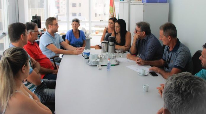 Mesa de reunião branca com uma dúzia de pessoas sentadas à volta conversando; ao centro, garrafa térmica e xícaras