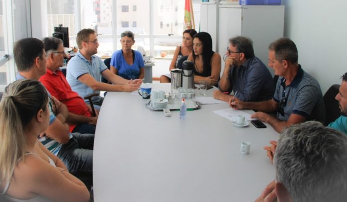 Mesa de reunião branca com uma dúzia de pessoas sentadas à volta conversando; ao centro, garrafa térmica e xícaras