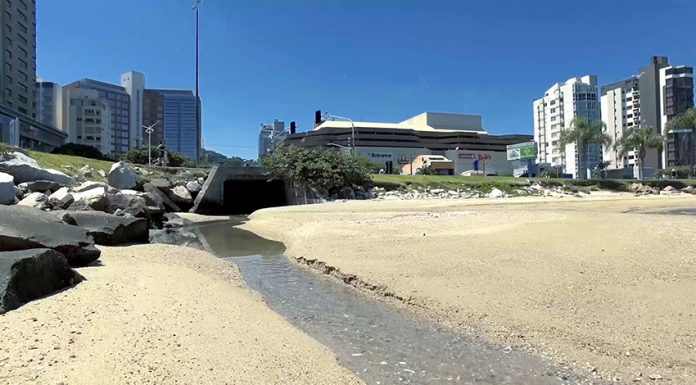 Um pequeno riacho de esgoto corre de uma vala cortando a areia da praia com prédios ao fundo e o céu azul