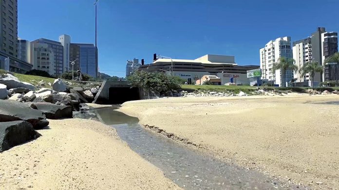 Um pequeno riacho de esgoto corre de uma vala cortando a areia da praia com prédios ao fundo e o céu azul