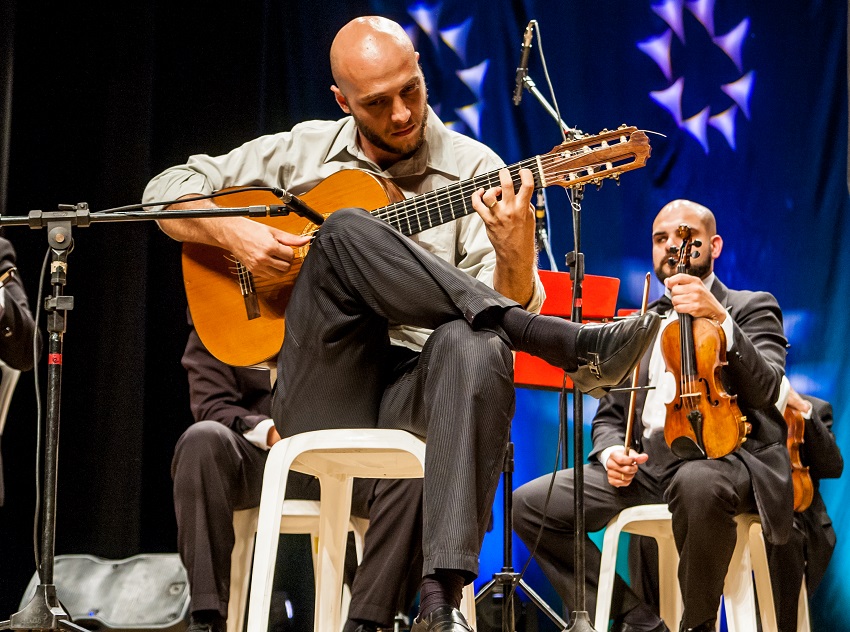 Homem careca toca violão, com as pernas cruzadas, sentado em um banquinho em palco com outro membro de orquestra ao fundo