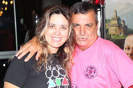 Mulher loira (à direita), usando camiseta preta, sorri para foto com homem (à esquerda) mandando beijinho e usando camisa rosa - o árbitro Margarida