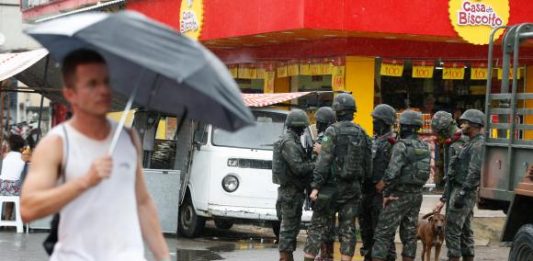 Pedestre segurando guarda-chuva passa por soldados militares conversando em uma rua de comércios