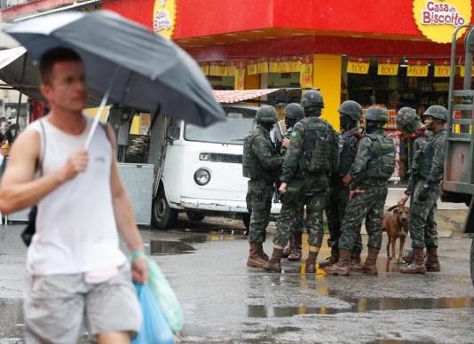 Pedestre segurando guarda-chuva passa por soldados militares conversando em uma rua de comércios