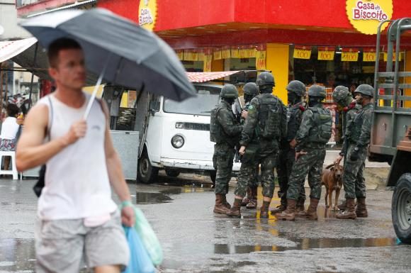 Pedestre segurando guarda-chuva passa por soldados militares conversando em uma rua de comércios