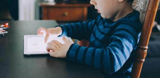 Pediatra alerta para exposição excessiva de jovens às telas Criança sentada em uma cadeira usa um tablet luminoso em cima de uma mesa