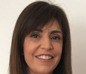 O mundo precisa de mais mulheres na liderança Imagem de uma mulher morena, sorridente, usando uma blusa preta e um colar