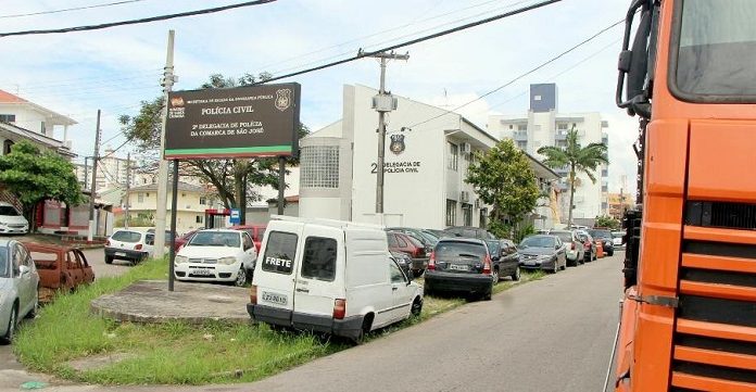 muitos carros largados em calçadas e ruas em volta de uma delegacia de polícia, colados uns nos outros