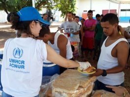 ONU e Governo instalam novo abrigo para venezuelanos em Boa Vista Mulher vista de costas, usando camiseta branca da ACNUR e boné azul entrega refeição a um homem, com um uma fila de venezuelanos atrás