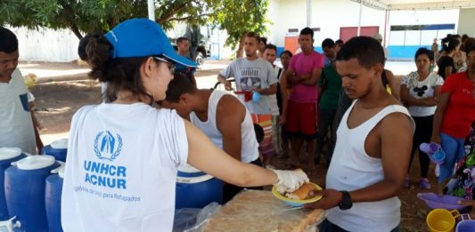 ONU e Governo instalam novo abrigo para venezuelanos em Boa Vista Mulher vista de costas, usando camiseta branca da ACNUR e boné azul entrega refeição a um homem, com um uma fila de venezuelanos atrás