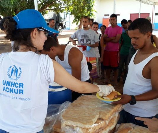 Mulher vista de costas, usando camiseta branca da ACNUR e boné azul entrega refeição a um homem, com um uma fila de venezuelanos atrás