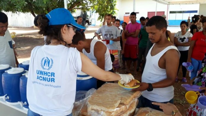 Mulher vista de costas, usando camiseta branca da ACNUR e boné azul entrega refeição a um homem, com um uma fila de venezuelanos atrás