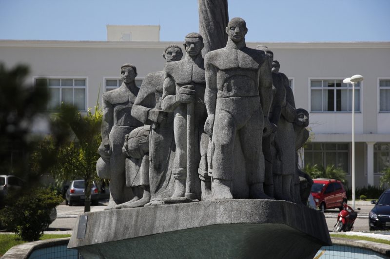 Monumento de pedra cinza em praça retratando imigrantes