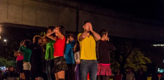 Grupo de atores em pé sobre uma mesa preta com as mãos tapando olhos, bocas e narizes