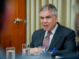 Empresário fala sentado na ponta de uma mesa com copos de água à frente