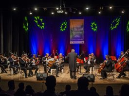 Orquestra se apresenta em um palco, com o maestro à frente, iluminada por uma luz azul de fundo