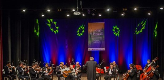 Orquestra se apresenta em um palco, com o maestro à frente, iluminada por uma luz azul de fundo
