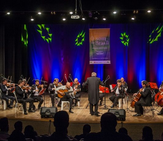 Orquestra se apresenta em um palco, com o maestro à frente, iluminada por uma luz azul de fundo