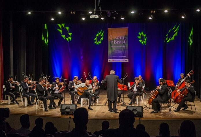 Orquestra se apresenta em um palco, com o maestro à frente, iluminada por uma luz azul de fundo