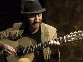 John Mueller apresenta novo álbum Homem usando chapéu preto e blazer bege olha e toca o violão em um close