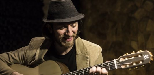 Homem usando chapéu preto e blazer bege olha e toca o violão em um close