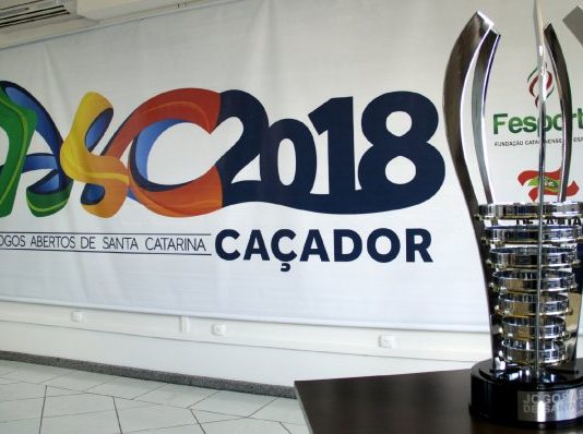 Imagem do troféu de campeão geral em primeiro plano, à direito, e ao fundo o cartaz oficial com os dizeres "Jasc 2018 Caçador"