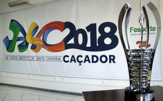 Imagem do troféu de campeão geral em primeiro plano, à direito, e ao fundo o cartaz oficial com os dizeres "Jasc 2018 Caçador"