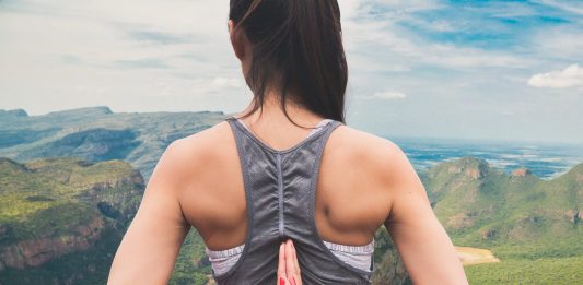 Imagem de uma mulher jovem de costas, com as mãos colocadas para trás em forma de oração; ao fundo, uma paisagem de montanhas