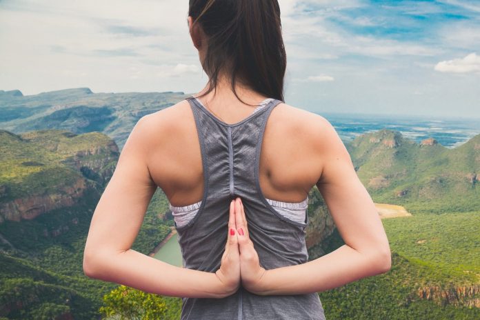 Imagem de uma mulher jovem de costas, com as mãos colocadas para trás em forma de oração; ao fundo, uma paisagem de montanhas