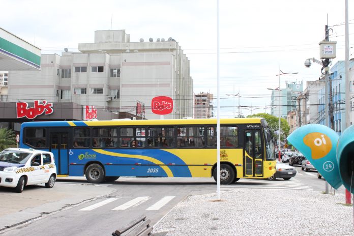Vista lateral de um ônibus coletivo azul e amarelo cruzando uma avenida com um calçadão no meio