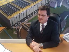 Zanotto sentado em uma mesa usando terno preto e óculo à frente de um painel com fotos da Casan