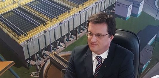 Adriano Zanotto assume gestão da Casan Zanotto sentado em uma mesa usando terno preto e óculo à frente de um painel com fotos da Casan