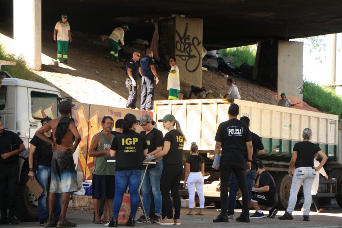 Foto em baixo do viaduto mostrando agentes de segurança na rua falando com usuários de drogas; ao fundo funcionários limpam e jogam lixo em caminhão