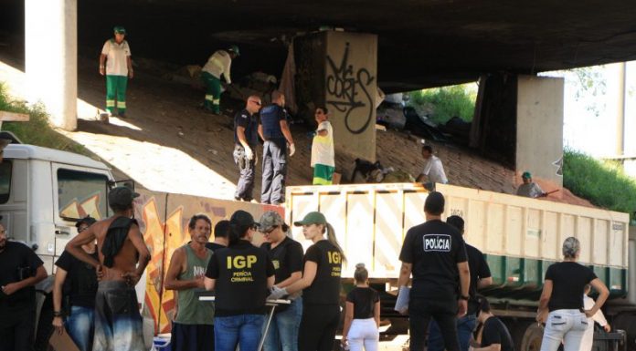 Foto em baixo do viaduto mostrando agentes de segurança na rua falando com usuários de drogas; ao fundo funcionários limpam e jogam lixo em caminhão