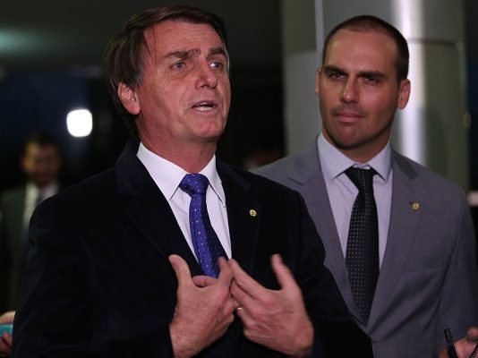Dois homens de terno dão entrevista; um, à esquerda, gesticula; o outro (o filho) fica olhando