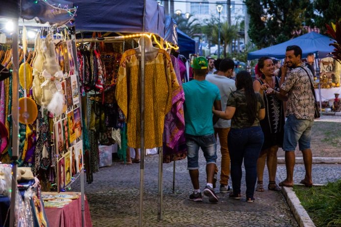 pessoas passam em frente à barracas de feiras com roupas à venda em uma praça no final do entardecer