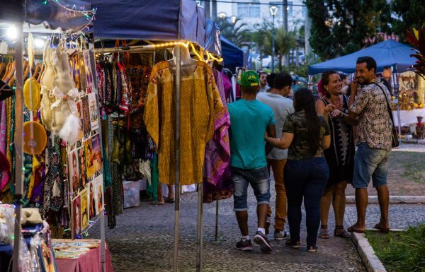 pessoas passam em frente à barracas de feiras com roupas à venda em uma praça no final do entardecer