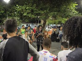 Garota negra lê uma poesia em seu celular de pé no centro de uma roda de espectadores numa praça