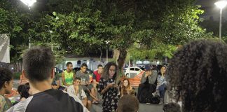 Garota negra lê uma poesia em seu celular de pé no centro de uma roda de espectadores numa praça