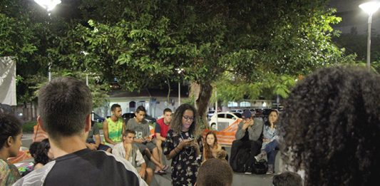 Slam Continente: A poesia está viva e batalhando na praça Garota negra lê uma poesia em seu celular de pé no centro de uma roda de espectadores numa praça