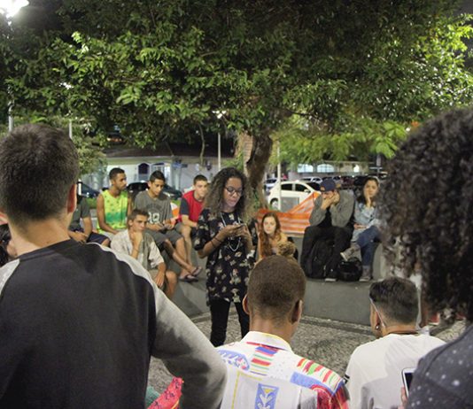 Garota negra lê uma poesia em seu celular de pé no centro de uma roda de espectadores numa praça