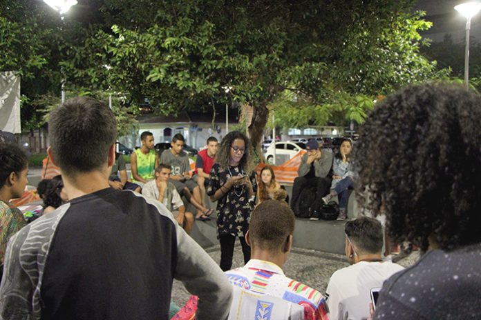 Garota negra lê uma poesia em seu celular de pé no centro de uma roda de espectadores numa praça