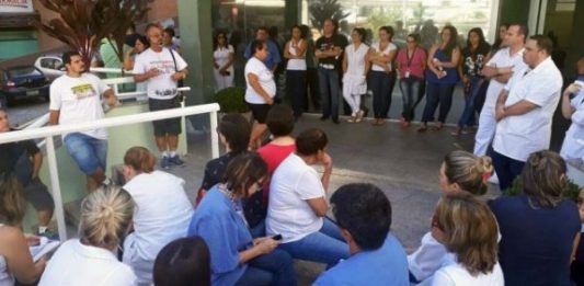 Saúde tem assembleia com indicativo de greve nesta quarta (4/4) Pessoas sentadas na escadaria em frente ao hospital ouvem um homem falar em pé