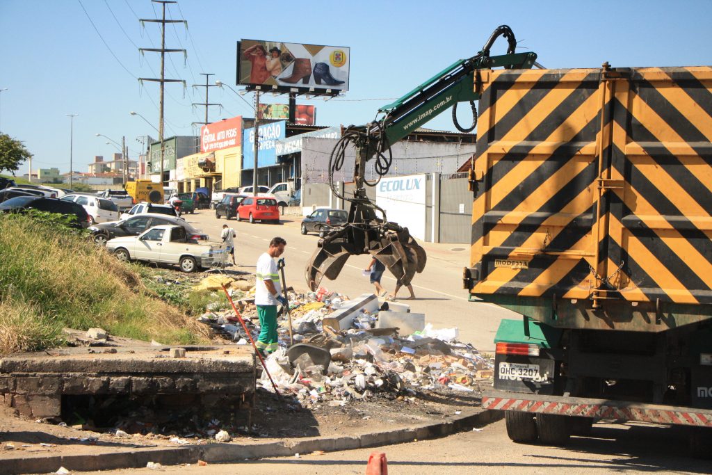 às margens de uma rua funcionário da comcap empurra uma montanha de lixo para que um braço mecânico de um caminhão possa pegar, estacionado ao lado
